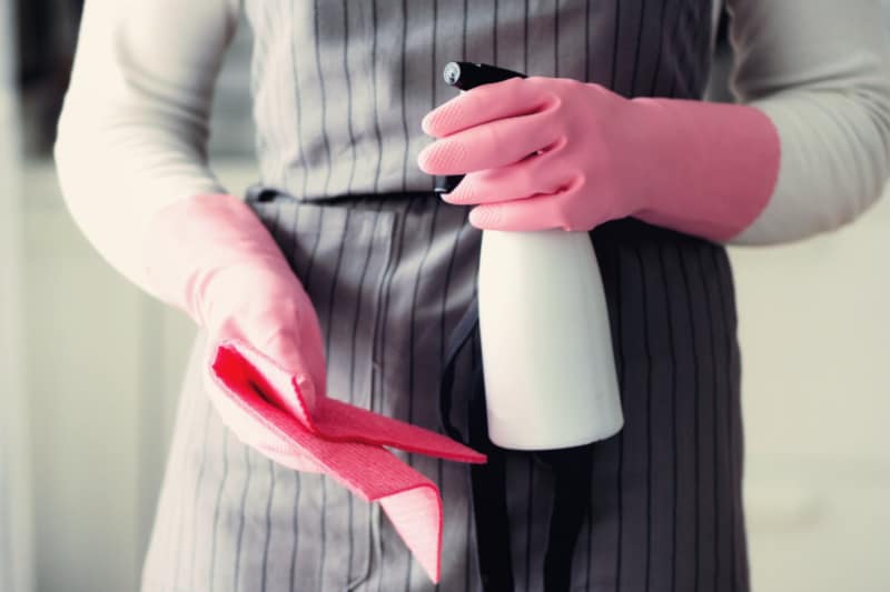 Mujer con guantes de goma y un delantal sosteniendo una botella de spray desinfectante casero y un paño de limpieza